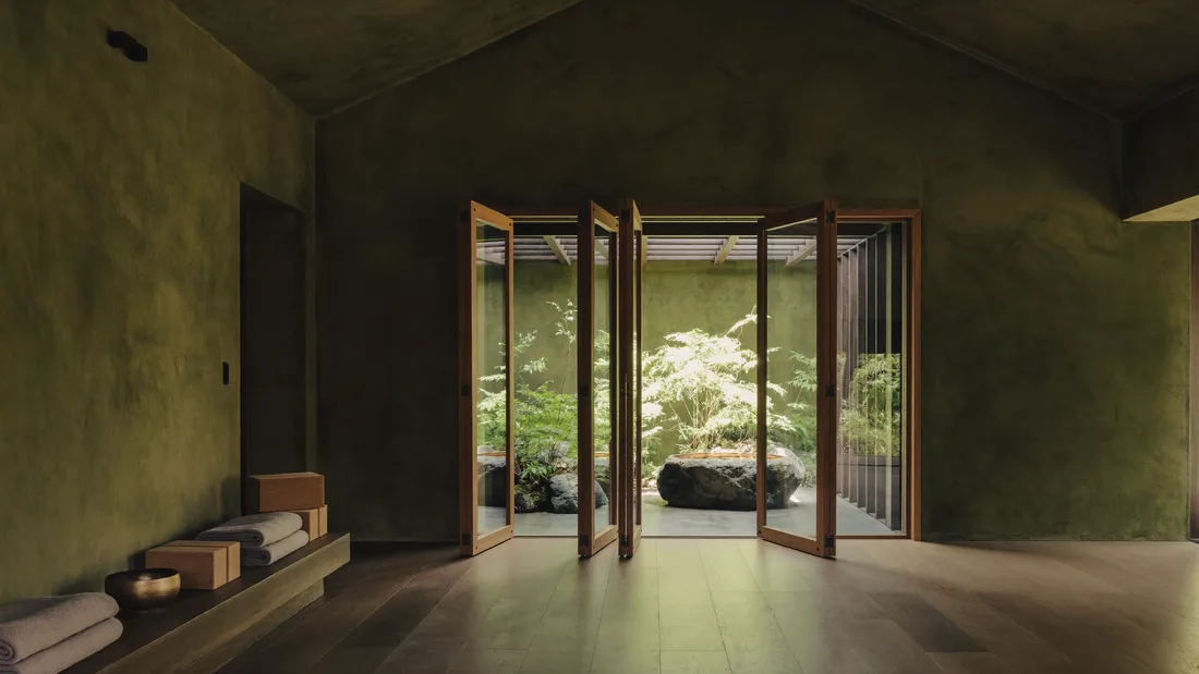 AI-generated wide view of a yoga shala with moss-green plaster walls, a teak floor and open wooden louvers onto a fern garden