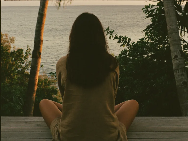 AI-generated view from behind of one person seated on a wooden deck at sunrise, facing the sea between palms