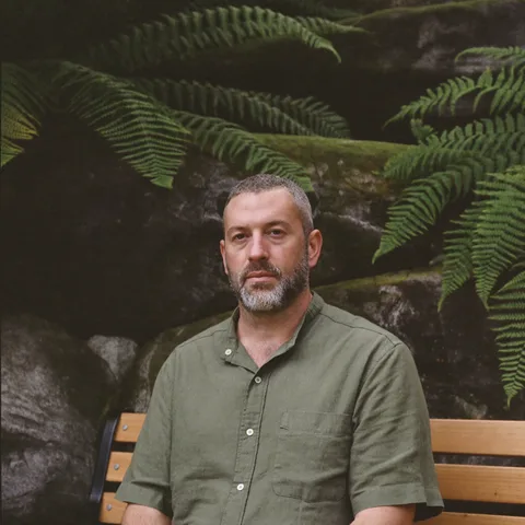 AI-generated portrait of Tomás, a fictional teacher, in an olive shirt on a bench in front of ferns